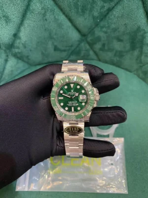 Black-gloved hand holding a Rolex Submariner "Starbucks" watch with green dial, green ceramic bezel, date window, and oyster bracelet featuring a "3135 CLEAN" sticker, with "CLEAN" packaging visible on a green background.