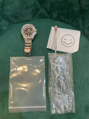 Black dial Rolex Submariner watch with protective plastic on bracelet, laid out on a green background alongside a smiling face polishing cloth, spring bar removal tool, and two clear zip-lock plastic bags.