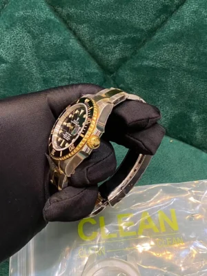 Black-gloved hand holding a two-tone Rolex Submariner watch with black dial, gold rotating bezel, and steel-gold bracelet, with a "CLEAN" labeled plastic bag nearby on a green background.