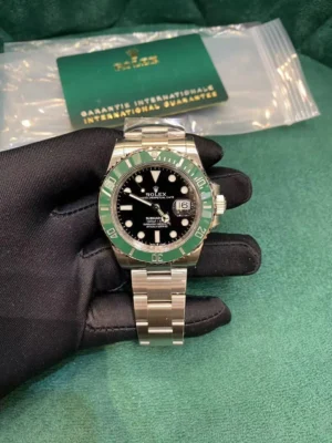 Black-gloved hand holding a Rolex Submariner "Hulk" watch with green ceramic bezel, black dial, date window, and oyster bracelet, next to a green Rolex international guarantee card in plastic packaging on a green background.