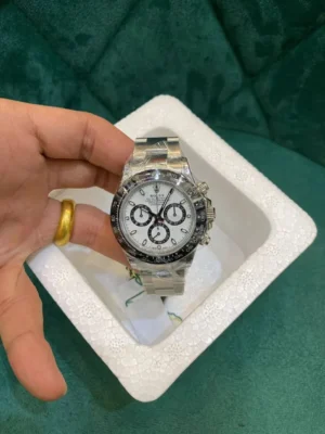 Hand holding a silver Rolex Daytona chronograph watch with white dial, black sub-dials, and black ceramic bezel, placed in a white foam tray on a green fabric background, with protective plastic on the bracelet.