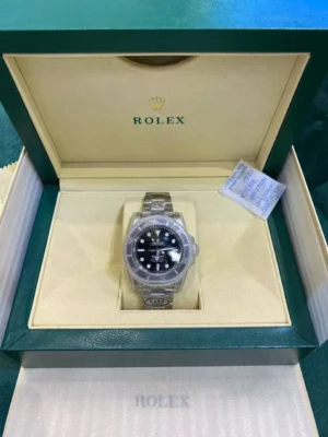 Black dial Rolex Submariner watch with protective plastic, resting in an open green Rolex presentation box with beige interior, silica gel packet, and warranty tag.